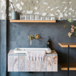 Area wastafel bergaya wabi-sabi atau industrial dengan dinding bertekstur abu-abu kasar, wastafel gantung bermotif batu dengan keran air kuningan di dinding, serta rak kayu minimalis di atasnya.