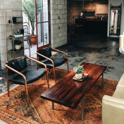 Ruang bersantai bergaya industrial mid-century dengan dinding batako ekspos, lantai beton gelap, karpet bermotif vintage, dua kursi kulit hitam berbingkai kayu, dan meja kopi kayu persegi panjang.